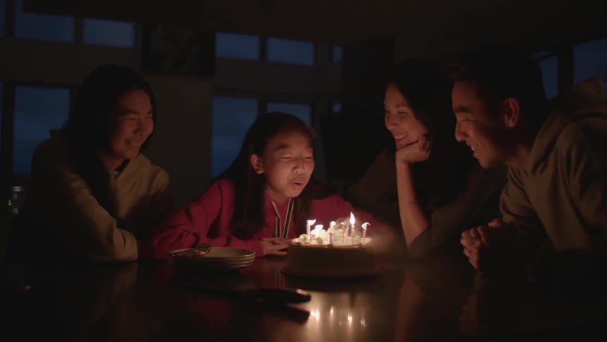 A child blows out the candles of her birthday cake with her family celebrating with her at home