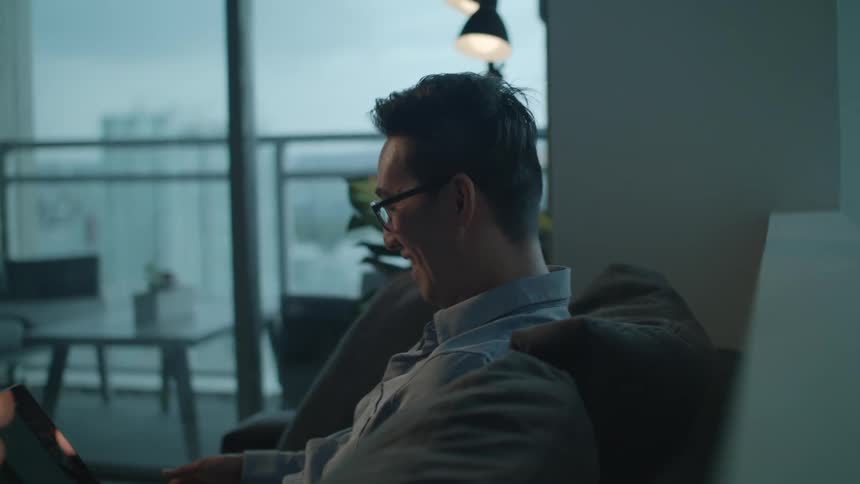 A man nods and laughs as he uses his laptop while sitting on a sofa in the living room of a house in the evening