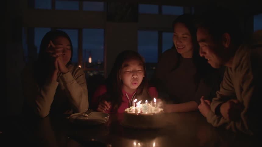 Family cheers daughter on as she blows out birthday candles leaving them in dark room