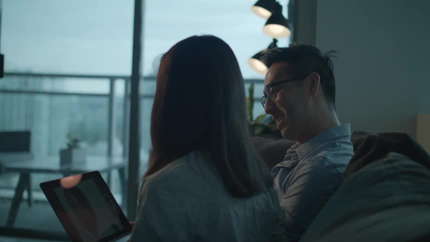 A man and a woman in a room sitting on a couch looking at a laptop with a wall and window in the background