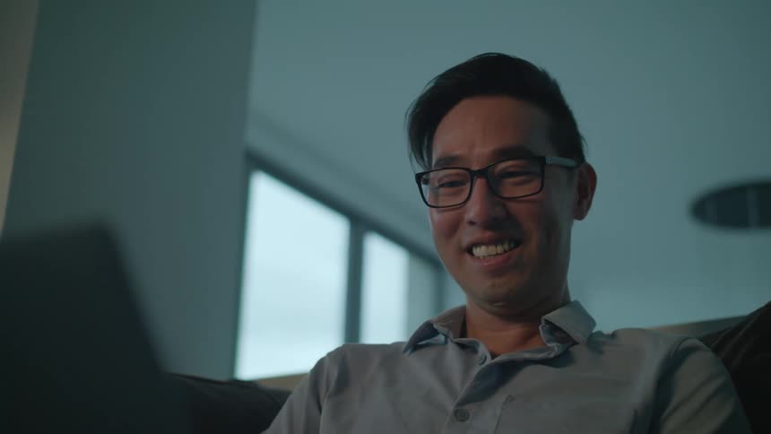 A man at home sits on a couch with his laptop and laughs and talks as he looks at it