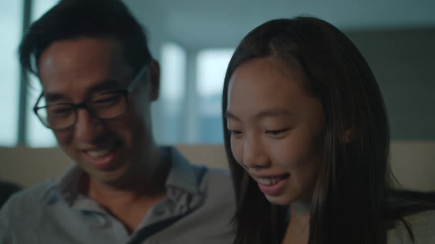 A male asian and female asian are sitting on a couch smiling and chatting