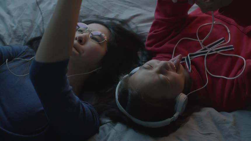 Sisters wearing earbuds and headphones take selfies resting in bed at home