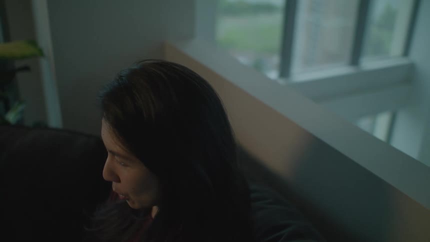 A woman in a room sitting on a couch with a plant a wall and window in the background