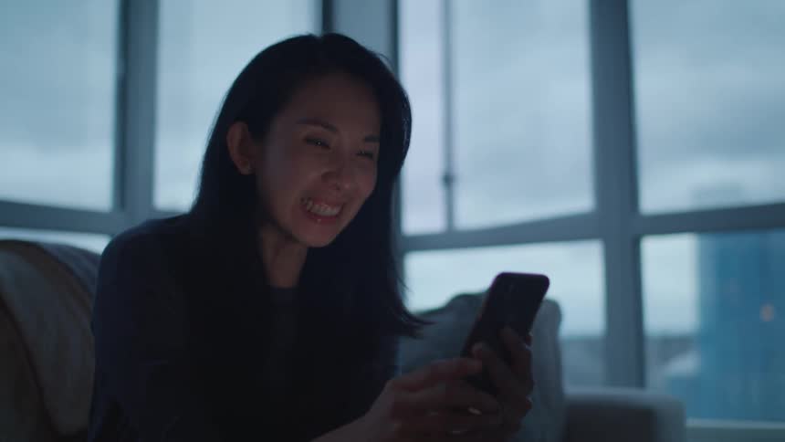 A woman in a room sitting on a couch looking at a smart phone with a wall and window in the background