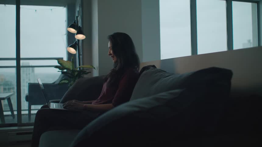 A woman in a room sitting on a couch using a laptop with a wall and window in the background