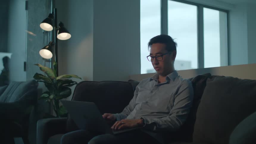 A man in a room sitting on a couch using a laptop with a wall and window in the background