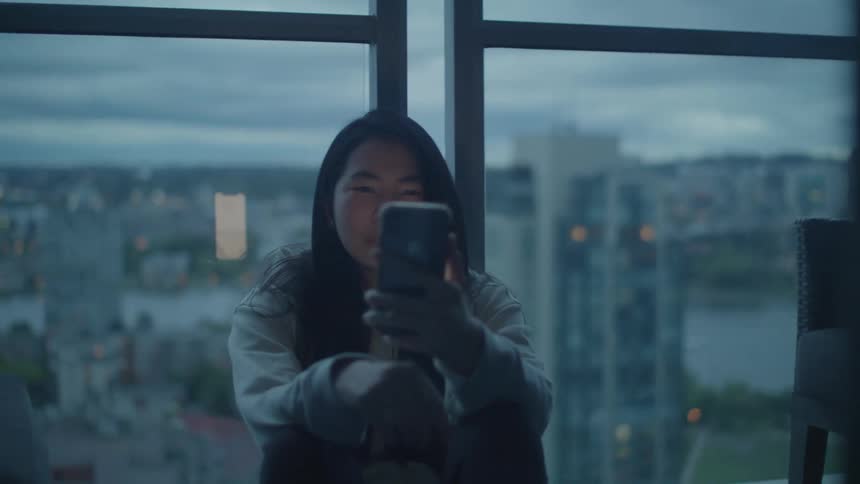 A teenage girl sits in front of a large picture window with the city skyline behind her and looks at her cell phone screen