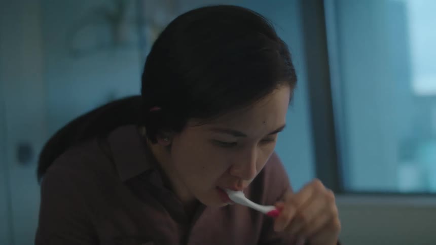 A woman checks her face in the mirror while brushing her teeth before spitting into the sink and rinsing her mouth