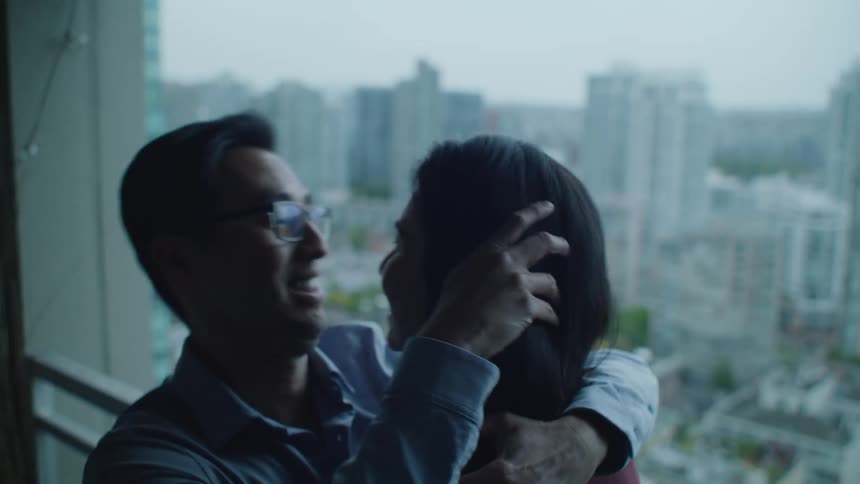 Couple kiss and smile standing in front of an apartment window in front of the city