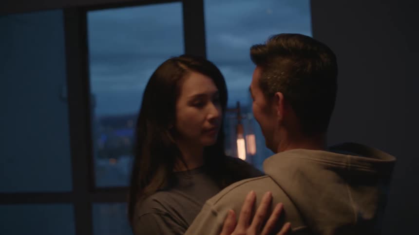 A man and a woman in a room hugging and kissing with a wall and window in the background