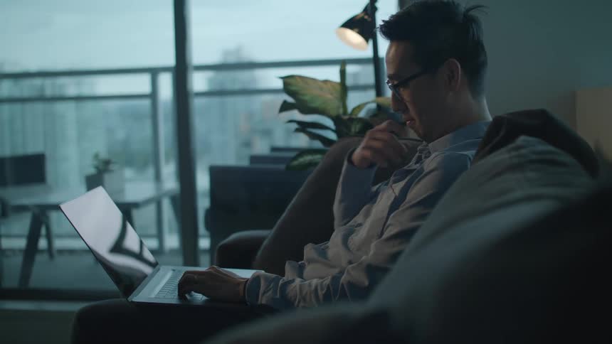 A man with glasses in a room using a laptop with a wall and window in the background