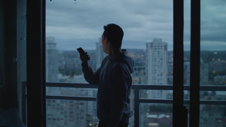 A man standing on a balcony participating in a video chat with his smartphone turns his phone to film the city for a moment before continuing his conversation