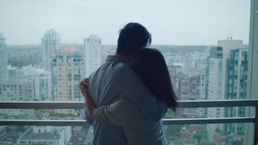 Woman with straight hair cuddles man wearing glasses and blue shirt watching the city from apartment balcony