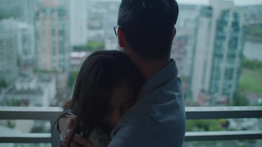 A man hugs a young woman before they look at one another and laugh on a balcony overlooking a city during the day