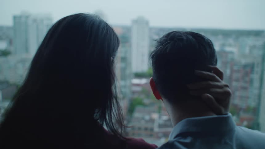 A man joins a woman on a balcony and the two look out over the city together