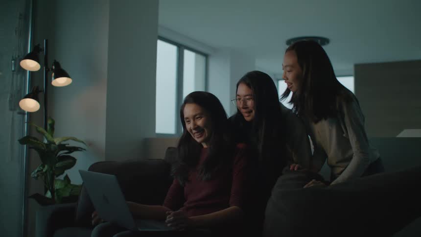 A mother and 2 daughters wave and talk into the camera on a laptop while inside of an apartment
