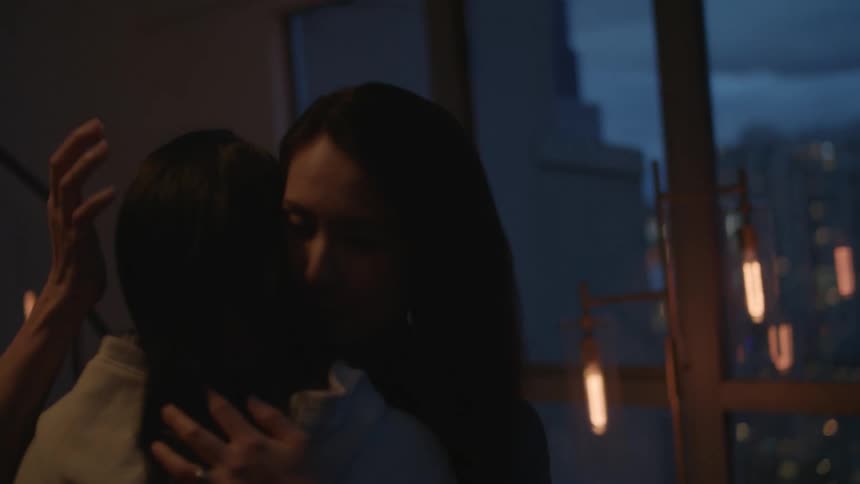 Woman with straight hair cuddles and kisses girl in dim lit apartment room