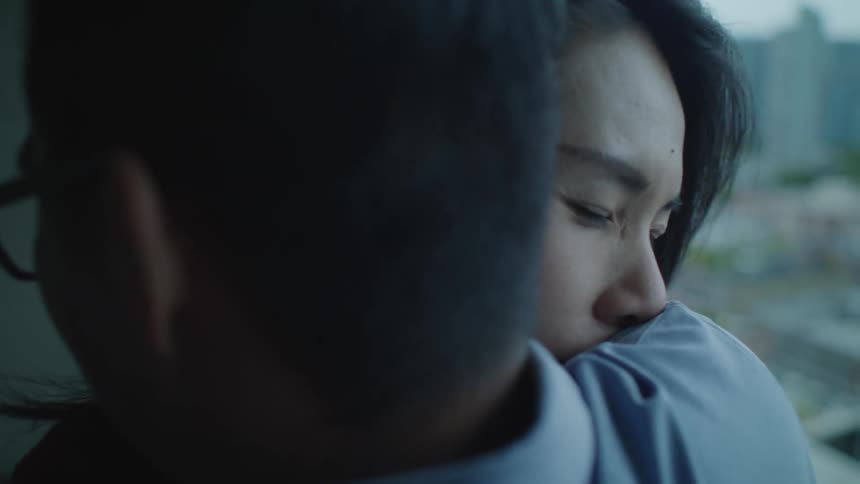 A man and woman hug and hold each other tightly while standing near windows overlooking the city