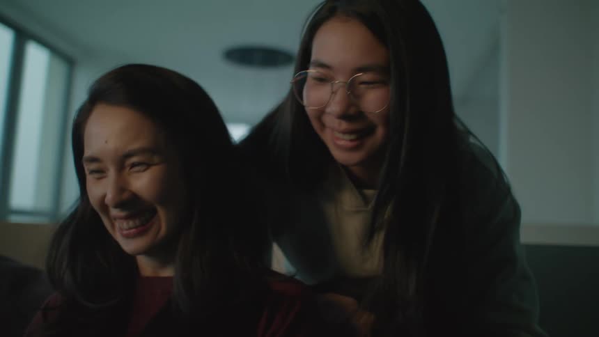 A daughter walks up to hug her mother who is watching something on a laptop