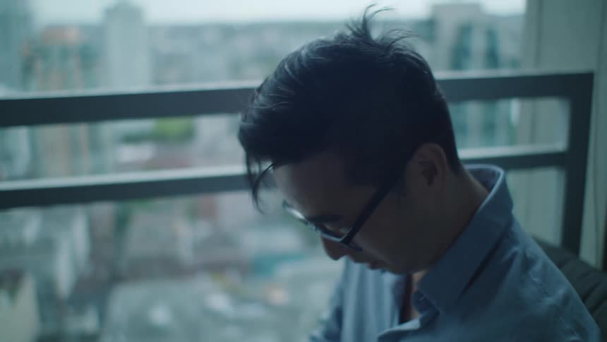 A man with glasses on a balcony with buildings the sky and clouds in the background
