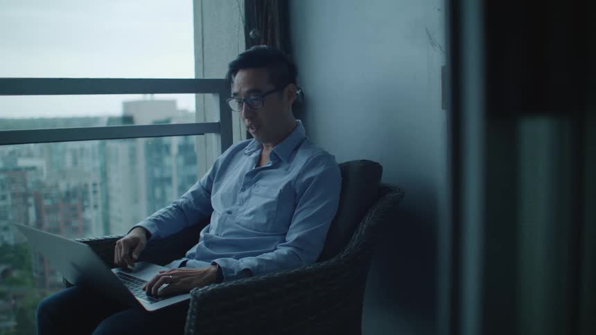 A man on a balcony sitting on a chair using a laptop with buildings the sky and clouds in the background