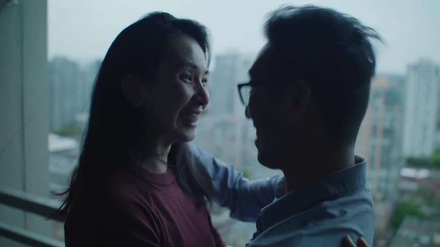 A man and woman are laughing and embracing in front of a window looking out over a city