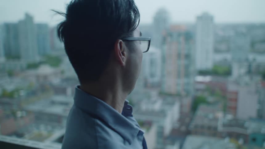 A man with glasses on a balcony with buildings the sky and clouds in the background