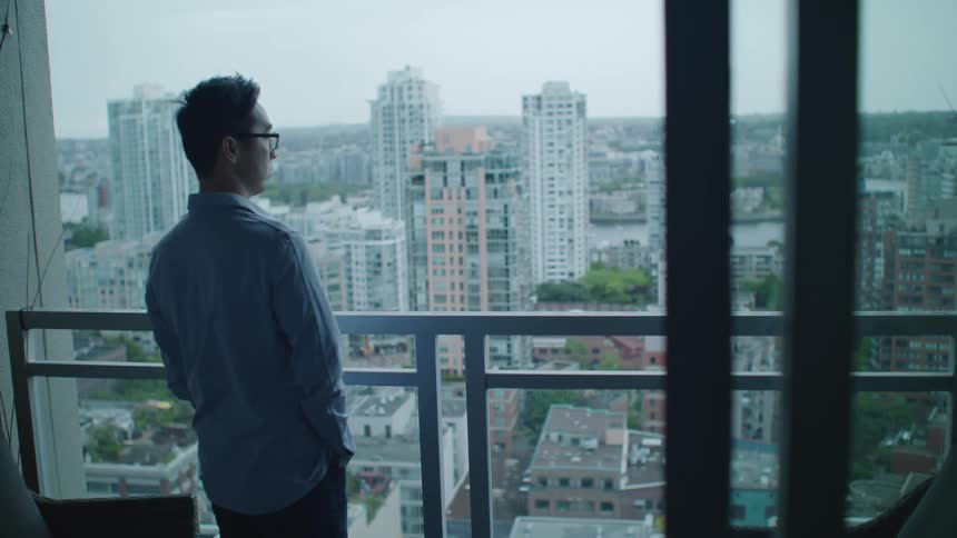 A man with glasses standing on a balcony with buildings the sky and clouds in the background