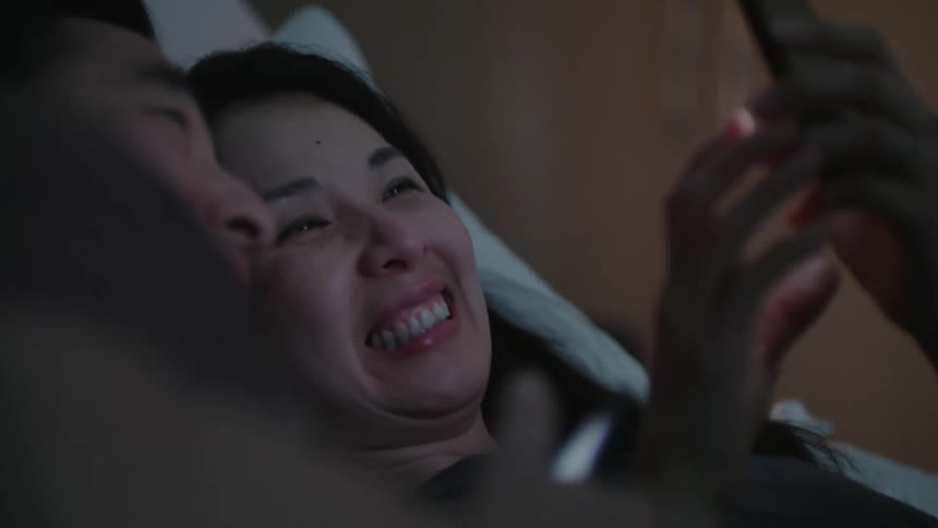 A woman and man laugh while lying side by side and looking at their smartphones