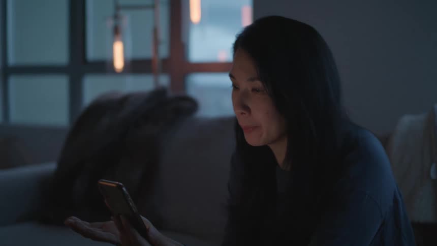 A woman in a room sitting on a couch looking at a smart phone with a wall and window in the background