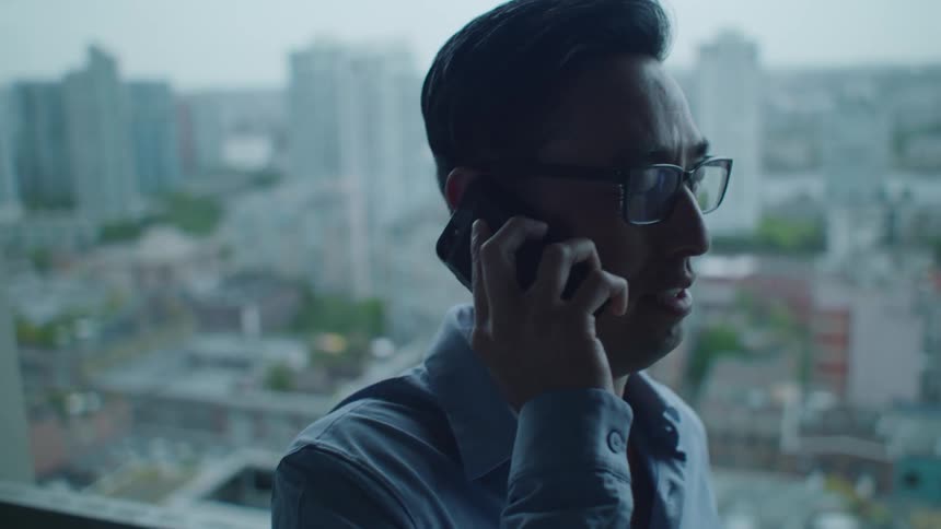 A man laughs as he talks on his cell phone in front of a large window showing a city