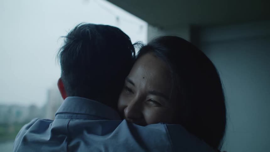 A man and woman laugh as they hug and kiss in front of a window