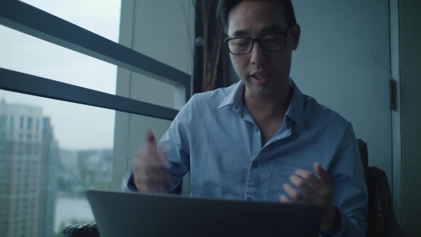 Young man wearing glasses talks while explaining in front of a laptop near a window