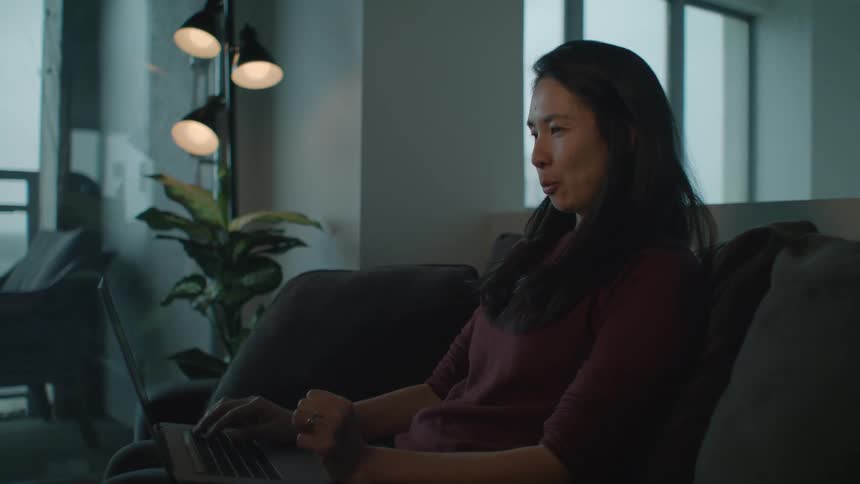 A woman sits on her couch at home with her laptop on her knee and laughs and talks as she video chats