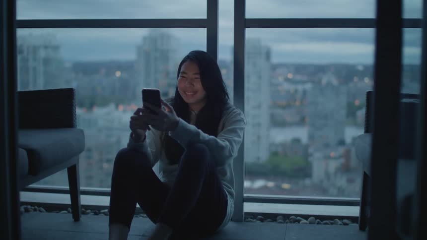 A teenage girl sits on the floor at home in front of a large window with the city skyline behind her and laughs as she looks at her cell phone