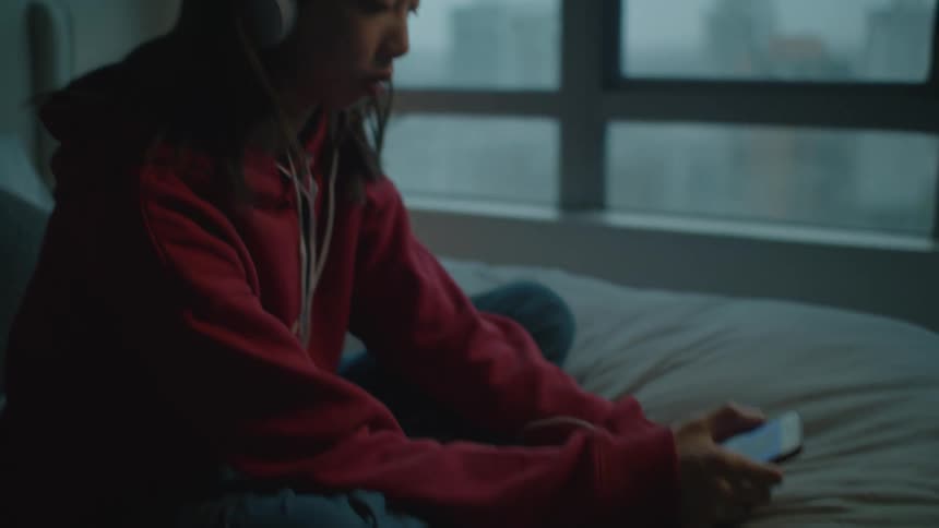 A young woman uses her smartphone to listen to music through her headphones while sitting crosslegged on a bed in a bedroom in the evening