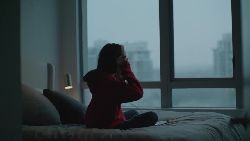 A girl in a room sitting on a bed with headphones listening to a smart phone with a wall and window in the background