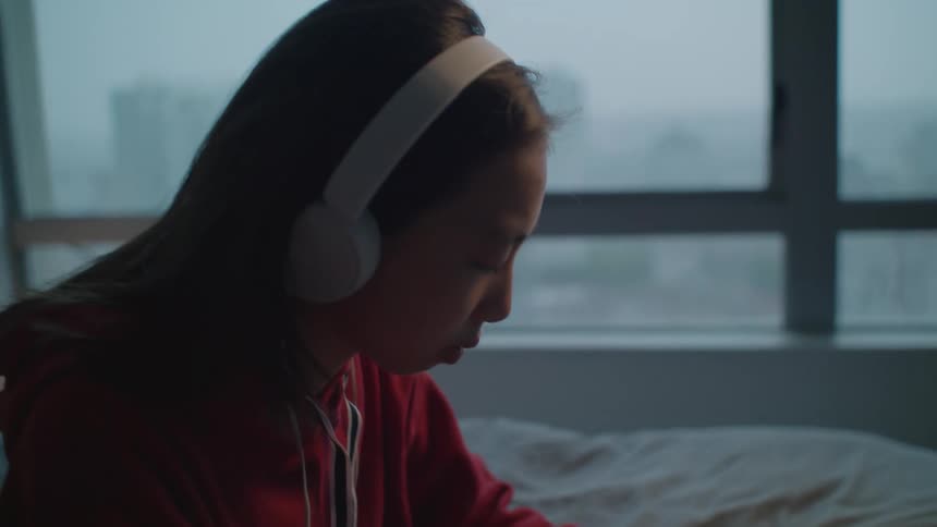 Teenage girl with red zip up and headphones sits on bed listening to music in her room
