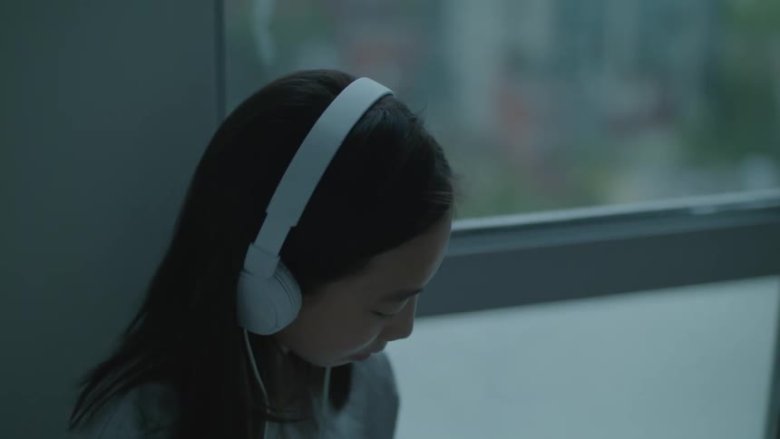 Young girl wearing headphones listens to music near a window at home