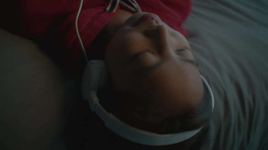 Young girl wearing headphones listening to music while laying in bed at home