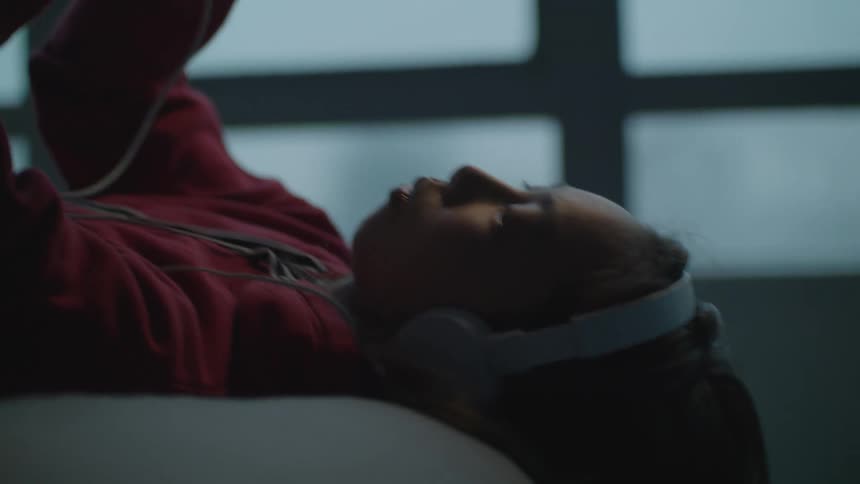 A girl in a room with headphones listening to a smart phone with a wall and window in the background