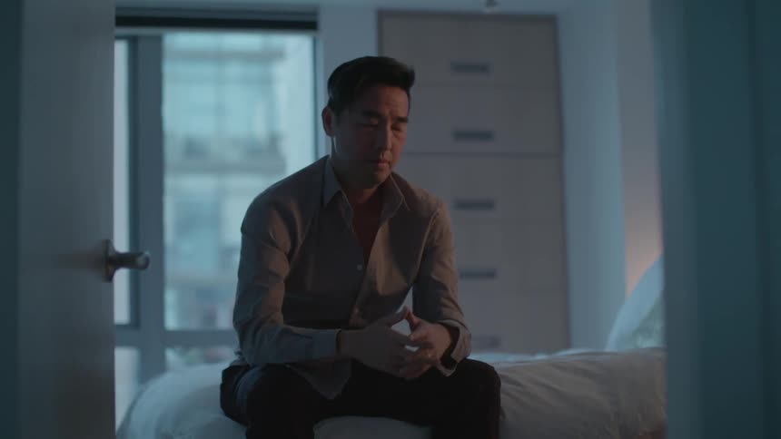 A man is sitting on a bed in an apartment during the day while thinking