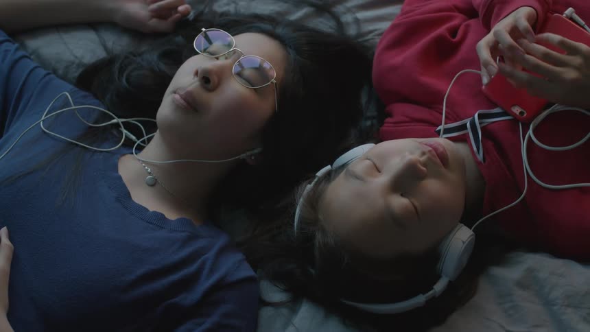 Two girls laying on a bed with headphones listening to smart phones