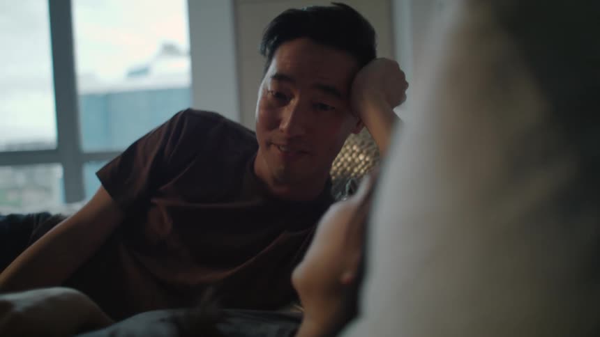 A man and a woman in a room laying on a bed with a wall and window in the background