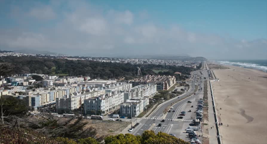 A road with vehicles next to buildings and the beach with the sky and clouds in the background