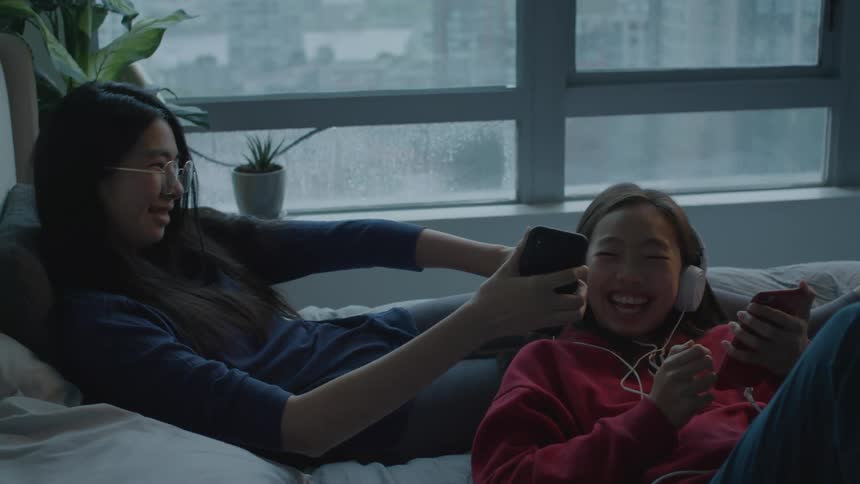 A young woman shows the screen of her smartphone to another girl resting next to her who laughs upon seeing it