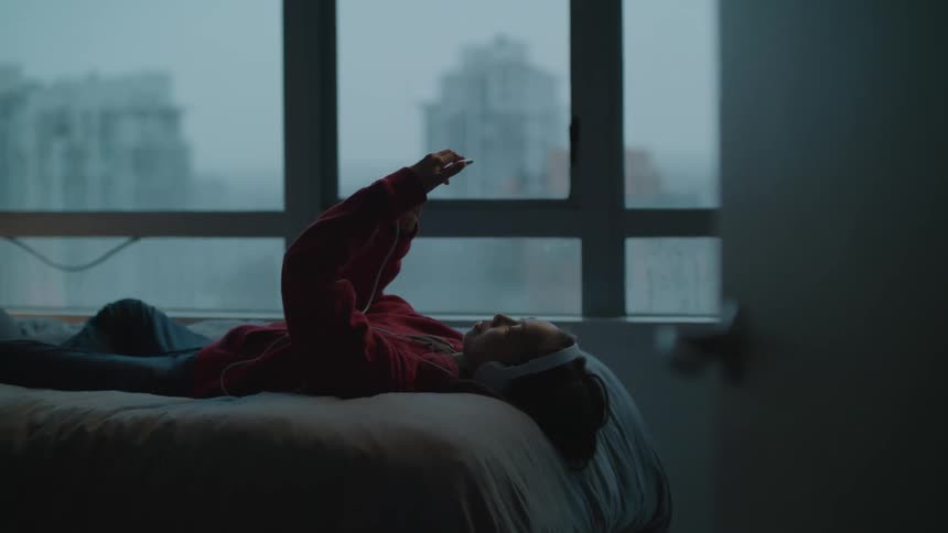 A girl in a room with headphones using a smart phone with a wall and window in the background