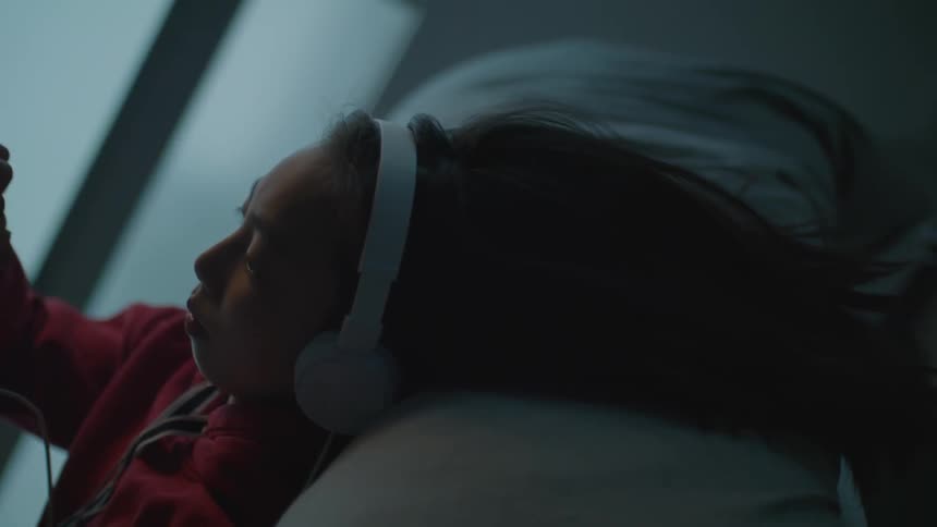 A young girl with headphones on leans back on a couch and uses her finger to touch a device
