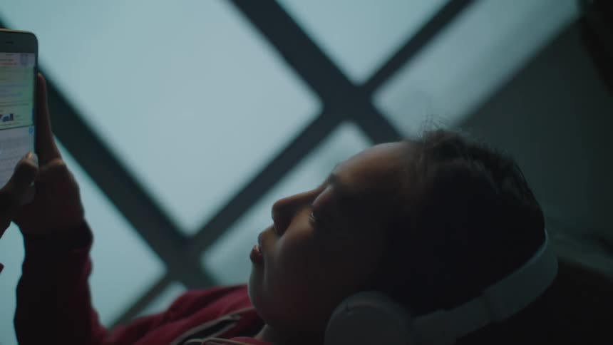 A woman is laying on a bed with headphones on while she looks at her cellphone